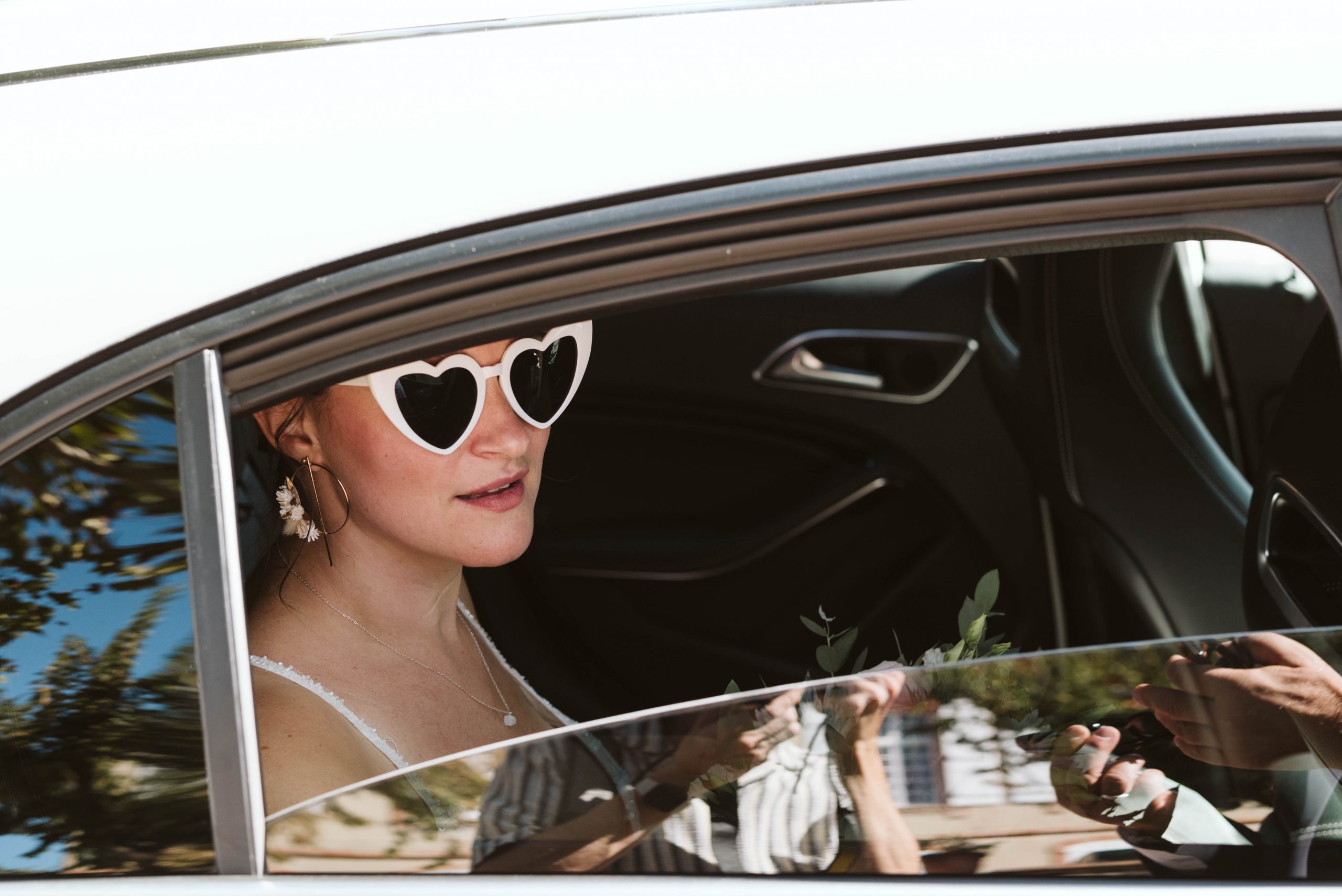 Portrait en noir et blanc d’une jeune femme portant des lunettes de soleil en forme de cœur, assise à l’arrière d’une voiture. La fenêtre entrouverte laisse apparaître son visage éclairé par la lumière naturelle, créant une ambiance rétro et cinématographique.