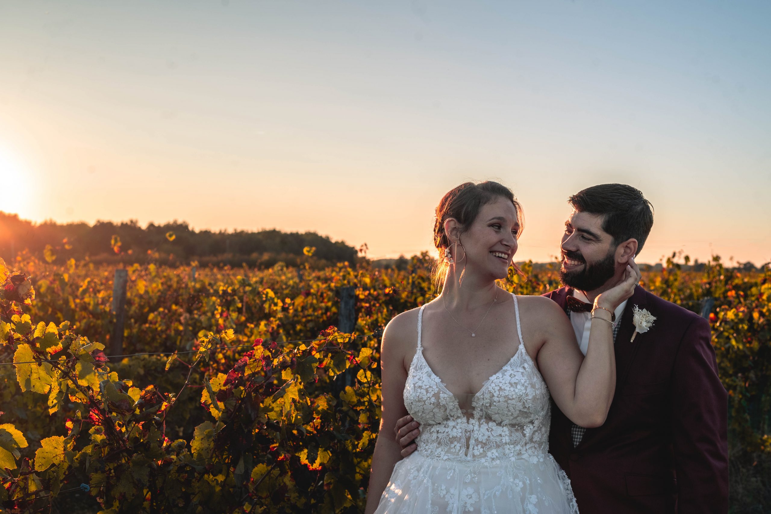 Couple de mariés souriants marchant dans les vignes au coucher du soleil, la mariée en robe blanche vaporeuse et le marié en costume bordeaux élégant, ambiance automnale dorée, photo de mariage lumineuse et naturelle en France.
