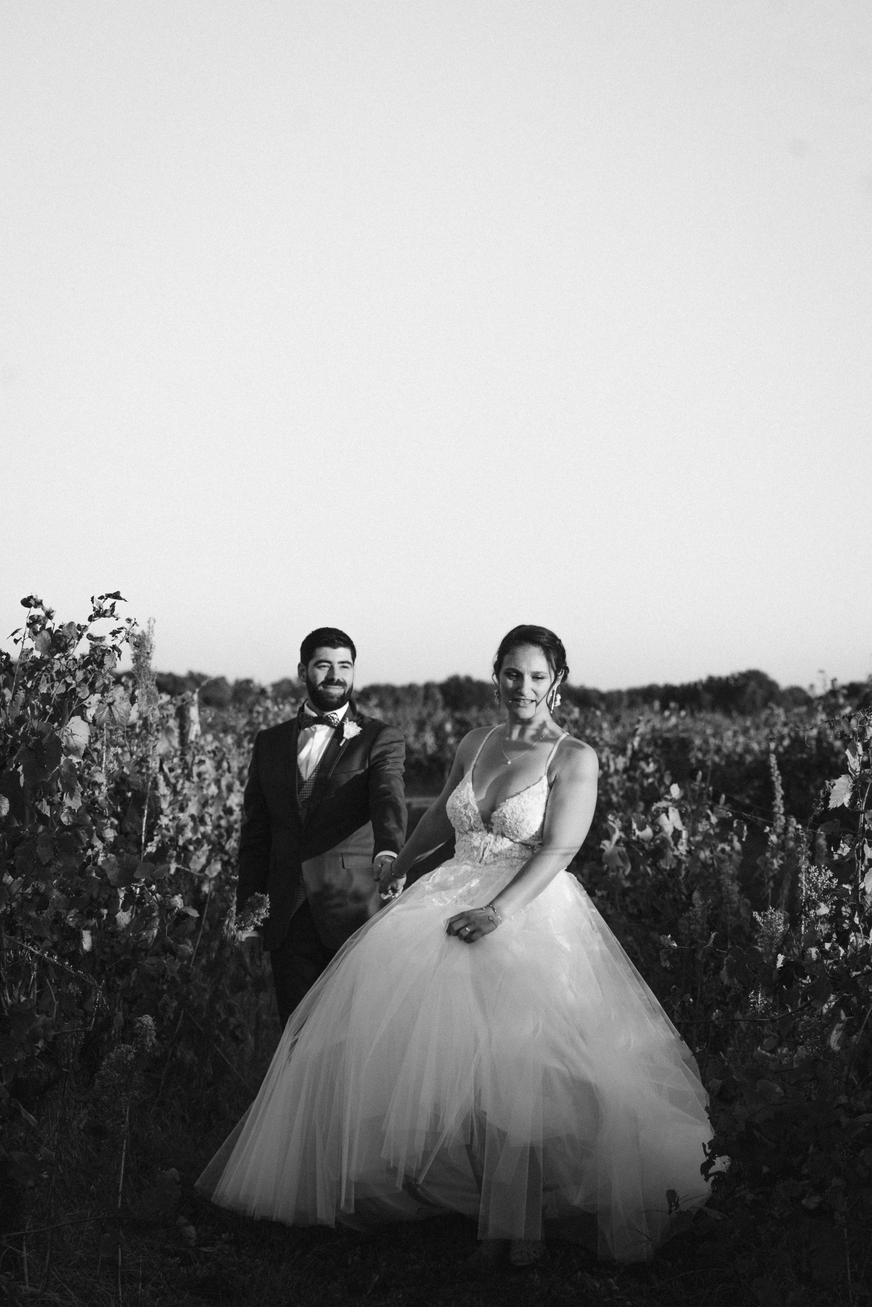 Couple de mariés souriants marchant dans les vignes au coucher du soleil, la photo est en noir et blanc