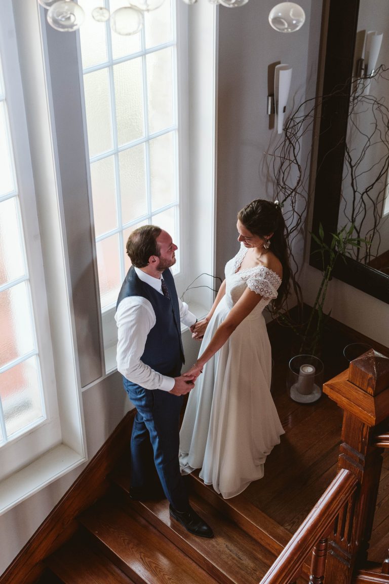 Photo lumineuse d’un couple de mariés se tenant les mains dans un escalier en bois, devant une grande fenêtre laissant entrer la lumière naturelle. La mariée porte une robe fluide à manches en dentelle et le marié un costume bleu marine élégant. Instant tendre et complice capturé lors des préparatifs de mariage.